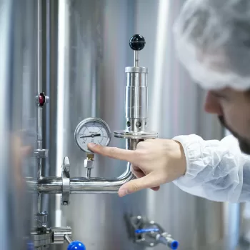 Técnico verificando medidor de vazão industrial em sistema de tubulação de aço inoxidável.